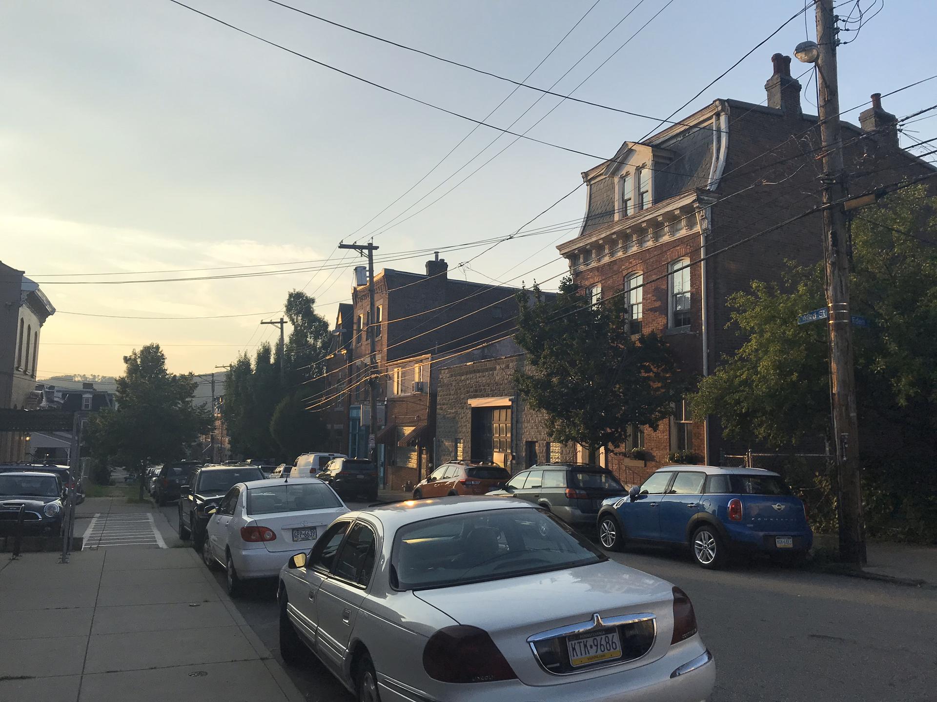 Residential street with parked cars and old buildings under an evening sky.