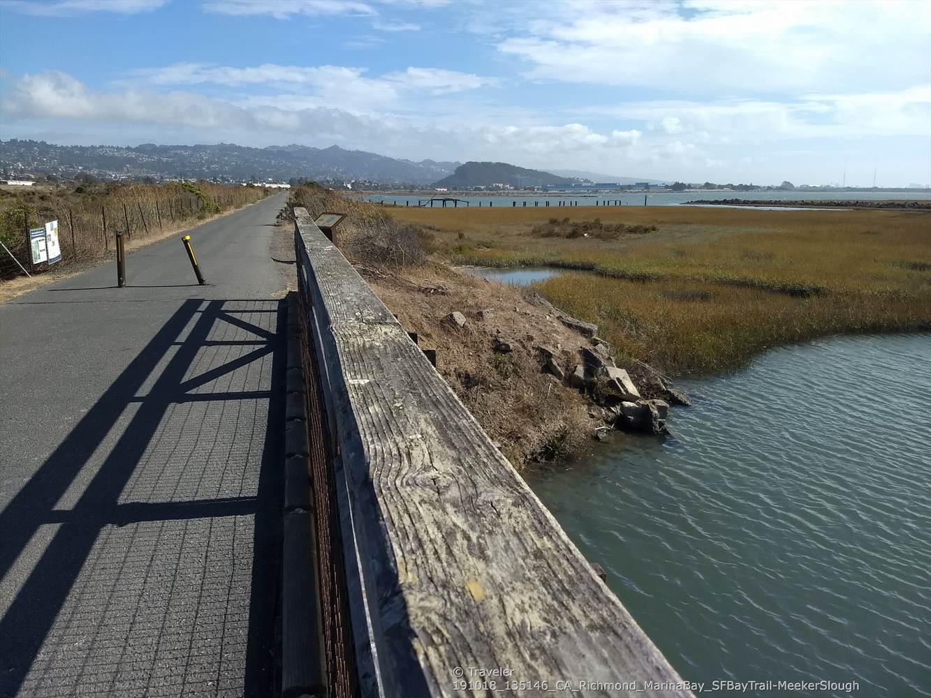 Paved path and wooden railing beside calm water and marsh under a bright sky.