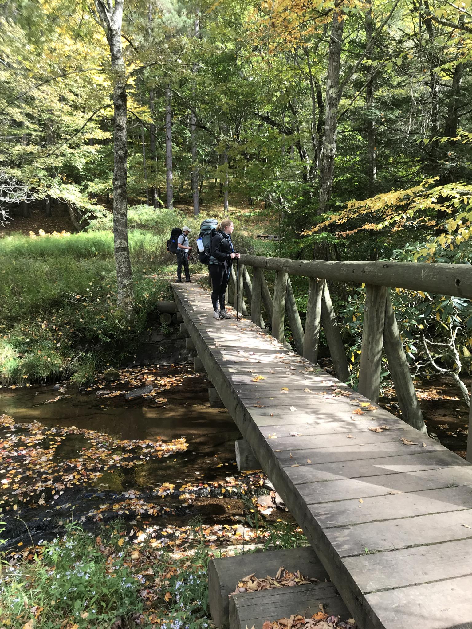 Two hikers with backpacks cross a wooden bridge over a stream in autumn.