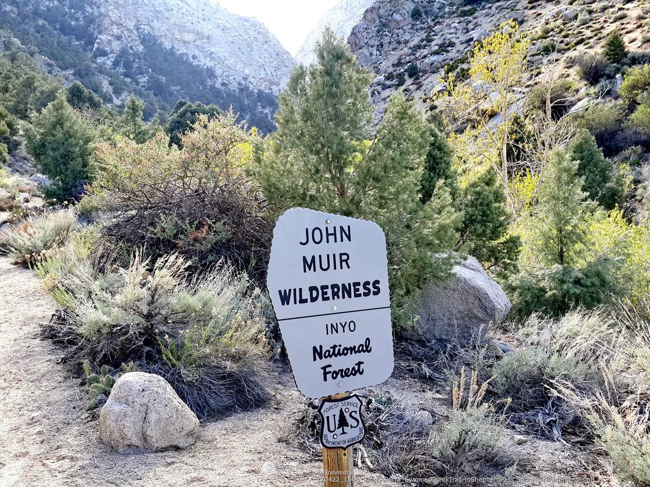 A wooden sign on a dirt path, surrounded by dry brush and mountains.