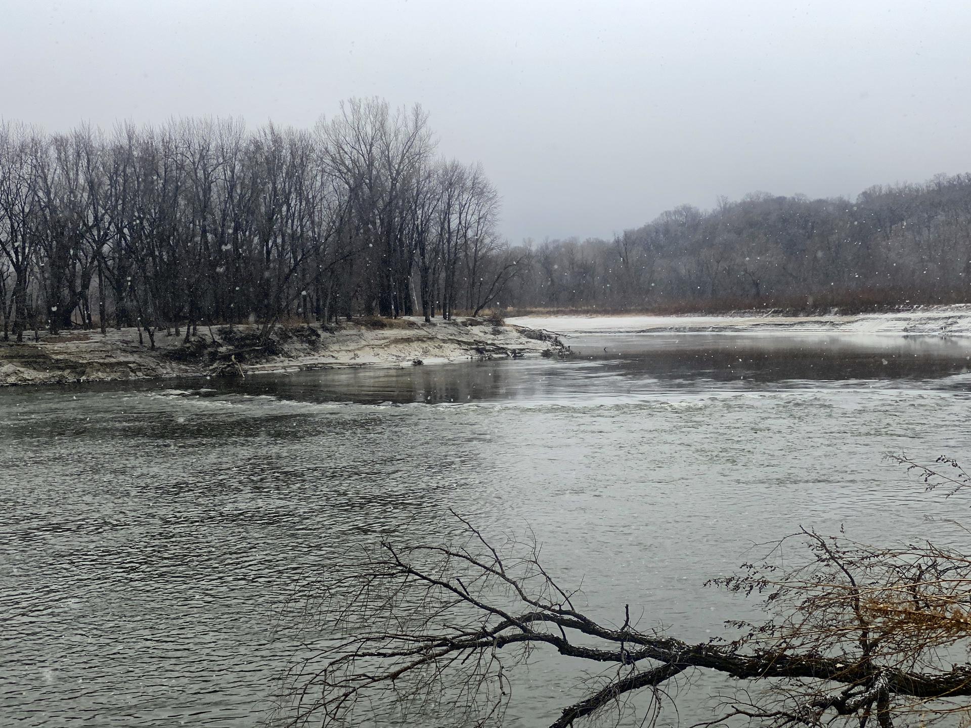 Winter river scene with bare trees, snow-covered banks, and falling snow.