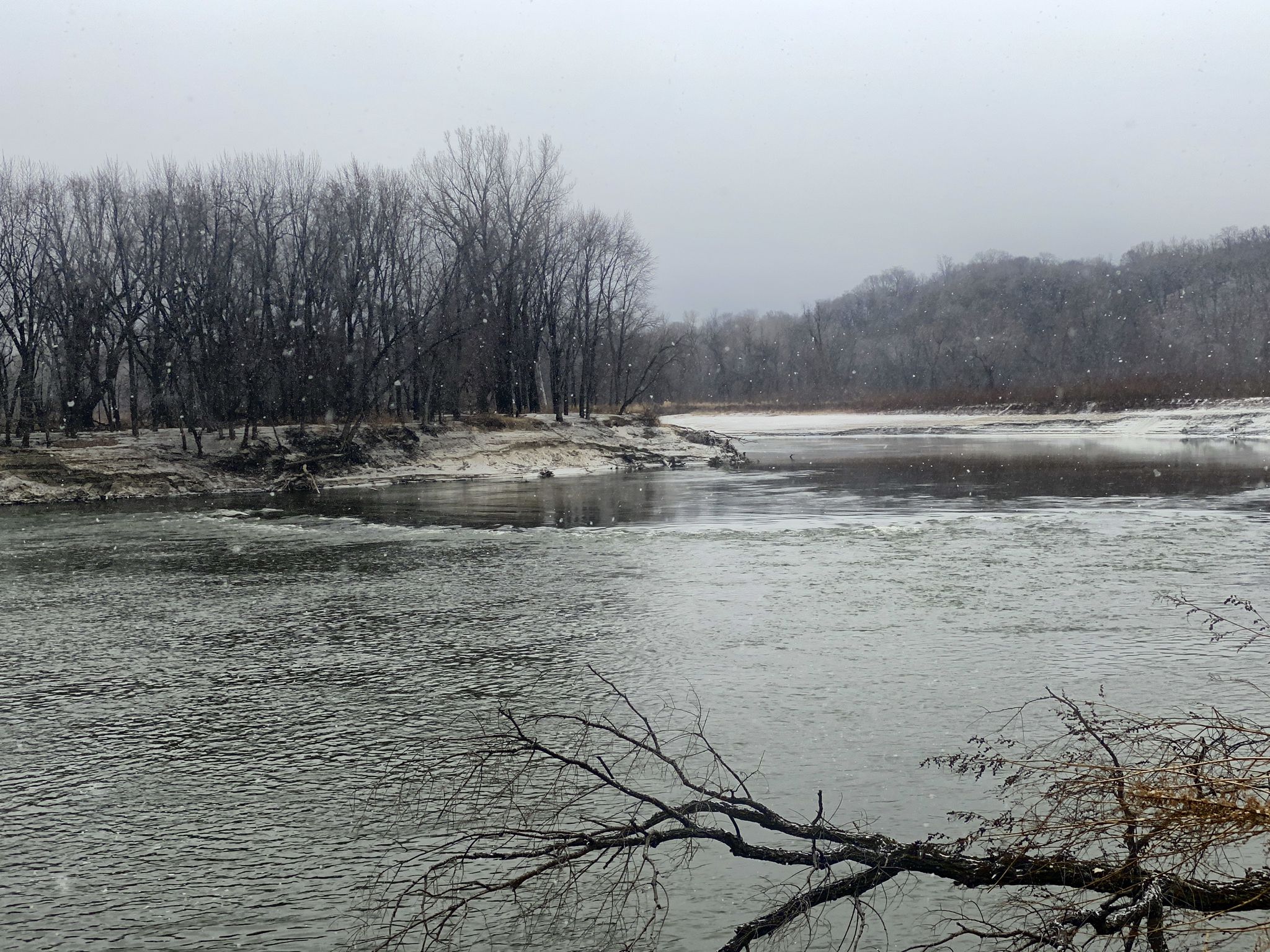 Winter river scene with bare trees, snow-covered banks, and falling snow. - Rapids Lake Trail