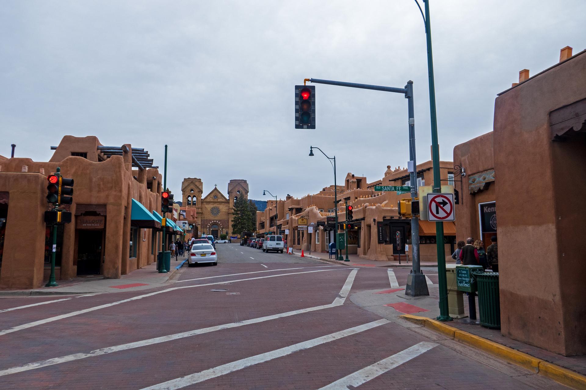 Urban street with adobe-style buildings, cars, and people under a cloudy sky.