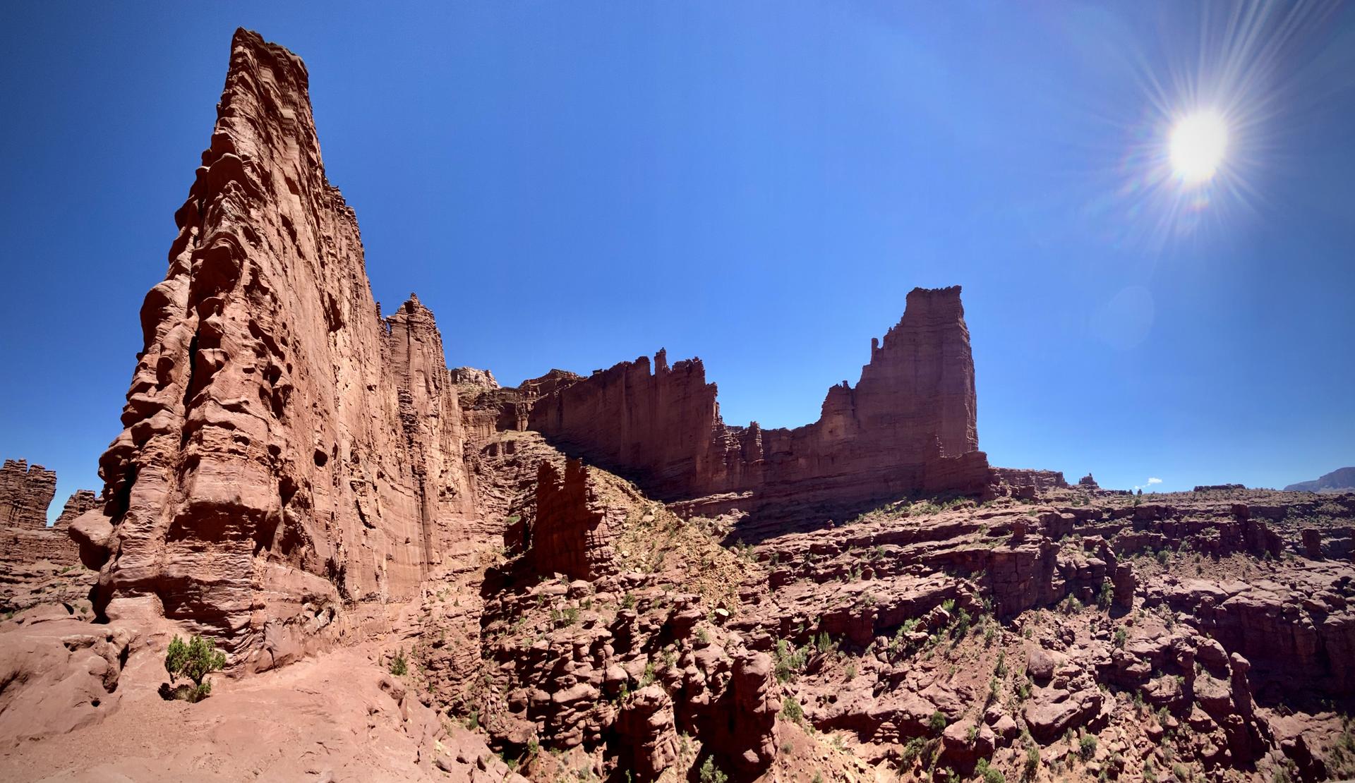 Dramatic low-angle view of a massive red rock spire and cliffs under a bright sun.