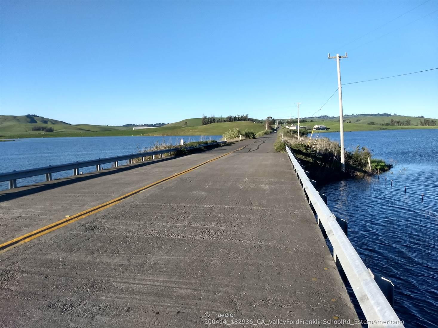 A sunny day shows a road crossing calm blue water with distant green hills.