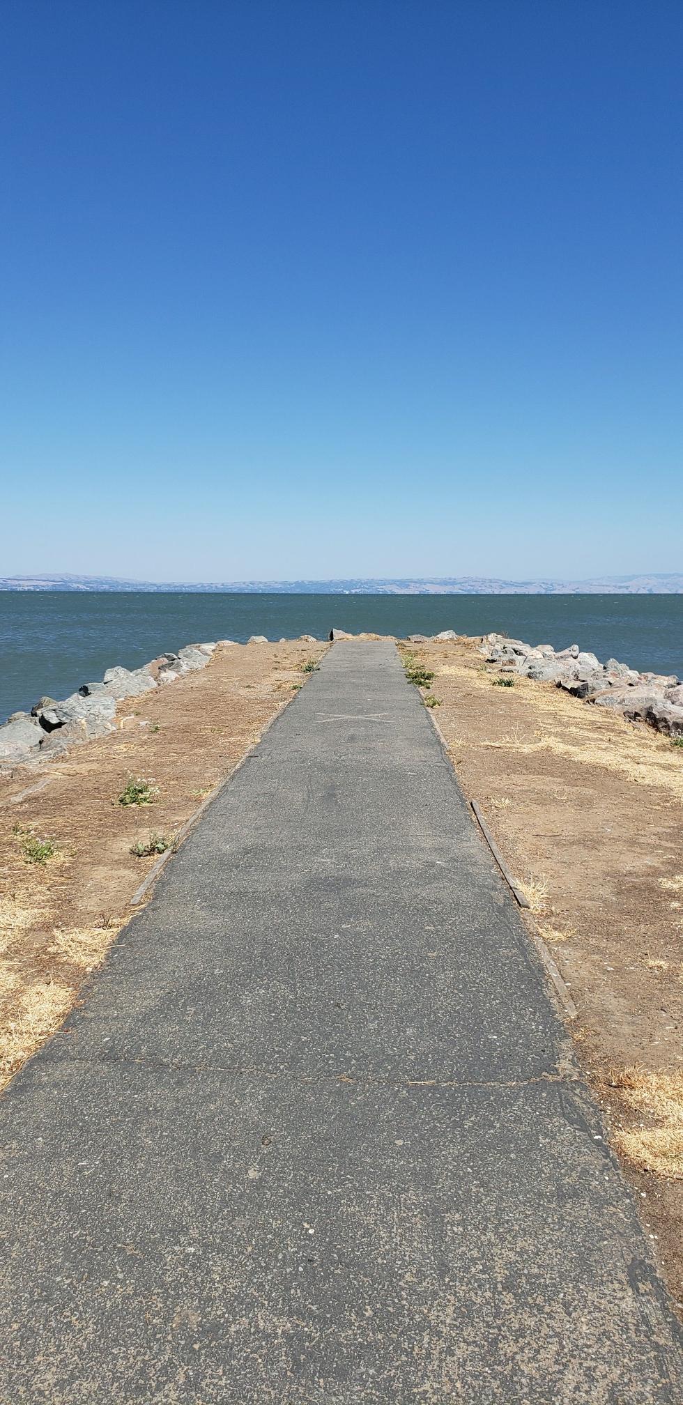 Narrow paved path extending into a wide body of water under a clear sky.