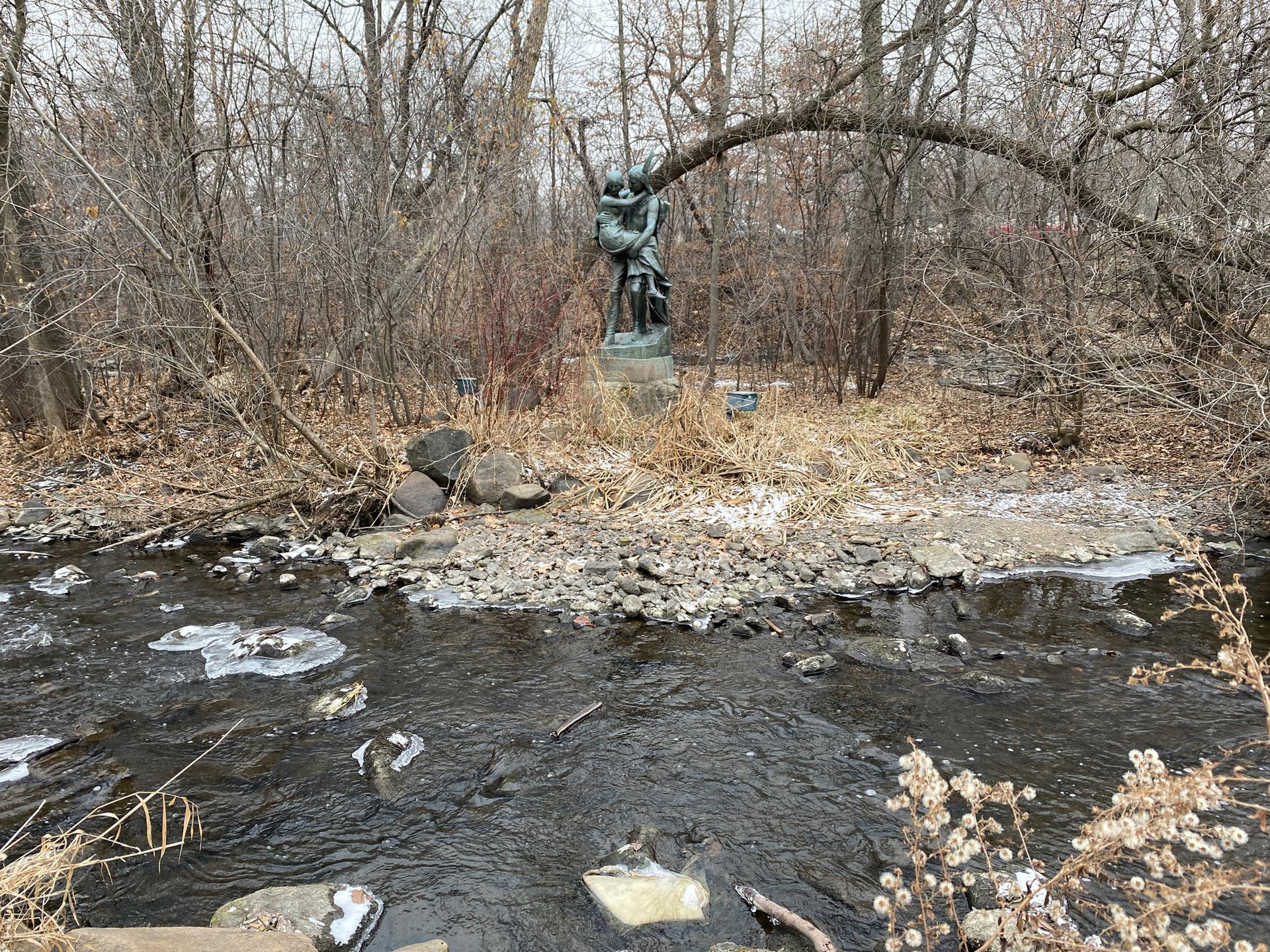 Bronze statue in a bare winter forest, overlooking a rocky stream with ice.