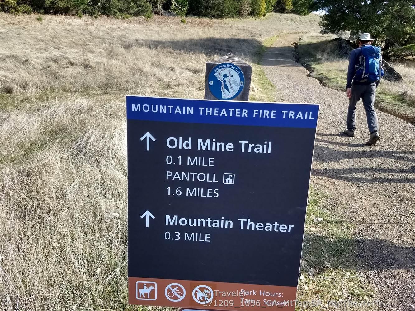 A person walks past a trail sign on a sunny, dry path.
