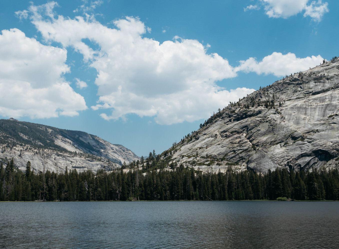 A calm lake with a forest and rocky mountains under a bright, cloudy sky.