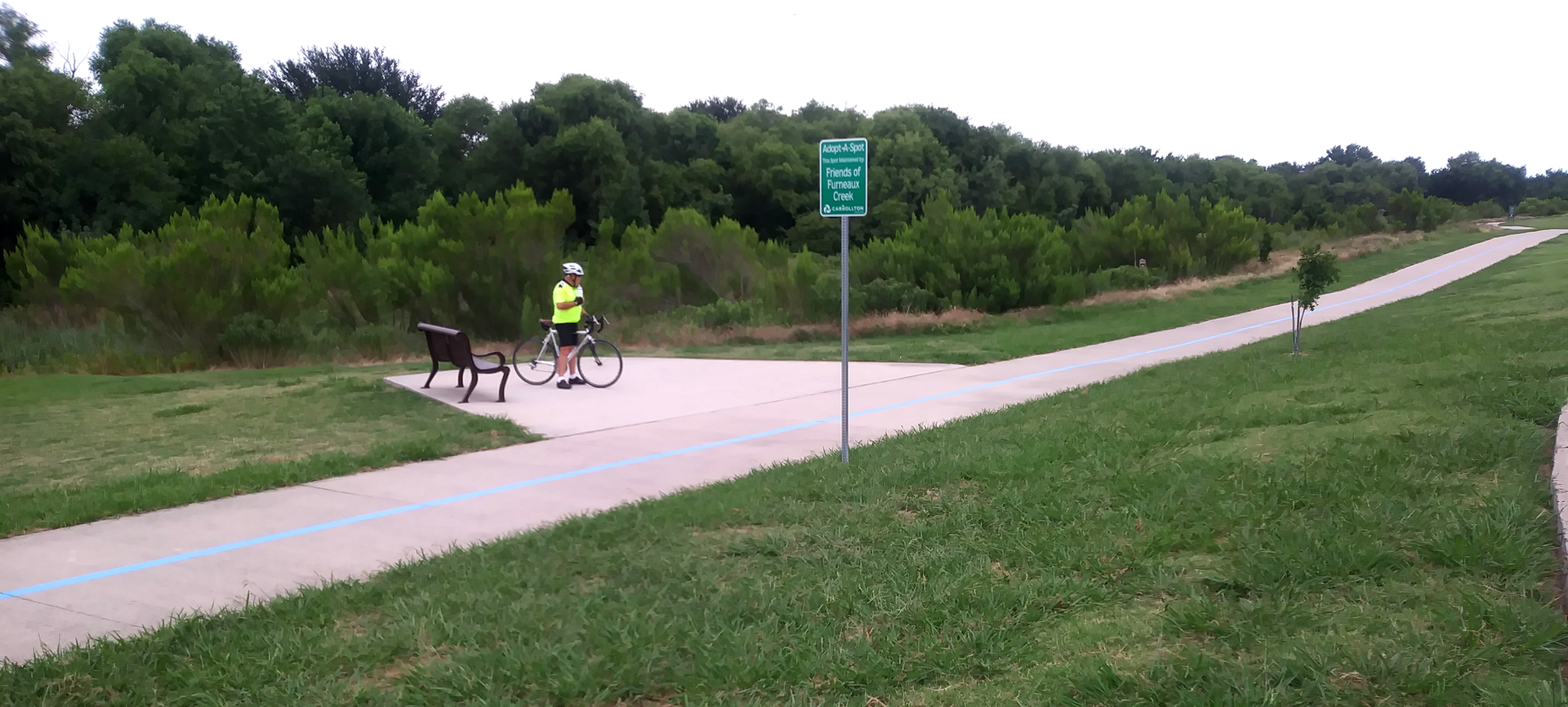 A person in a yellow shirt stands with a bicycle on a paved trail.