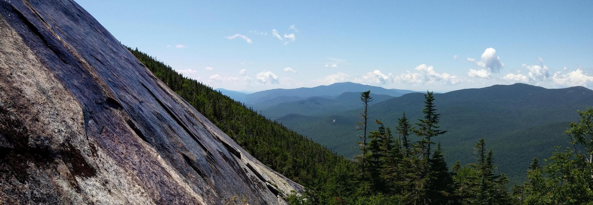 Steep rock face next to a forest, with distant mountains under a blue sky.