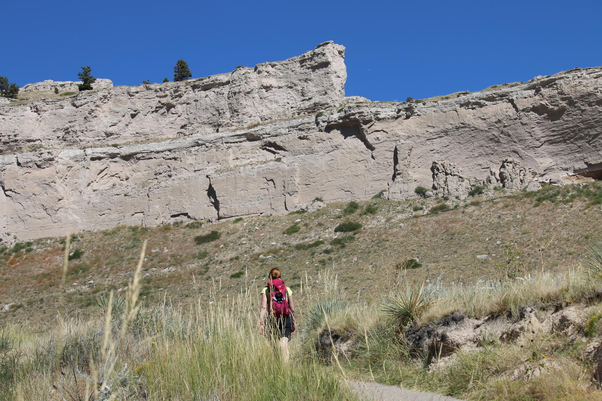 Hiker with a red backpack ascends a path towards a large rock formation.