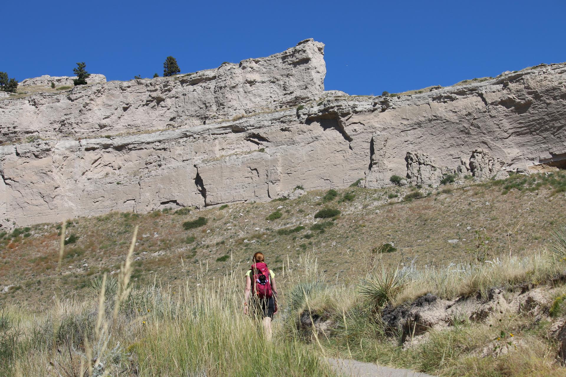 Hiker with a red backpack ascends a path towards a large rock formation.