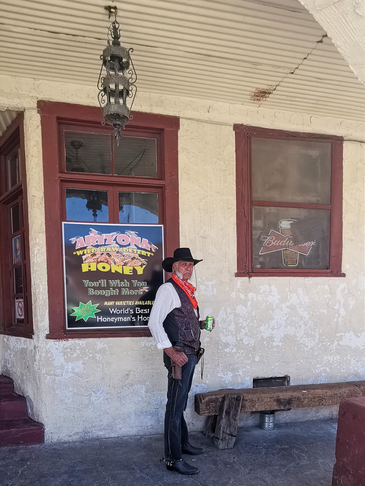 Man in cowboy attire holding a drink stands outside a building.