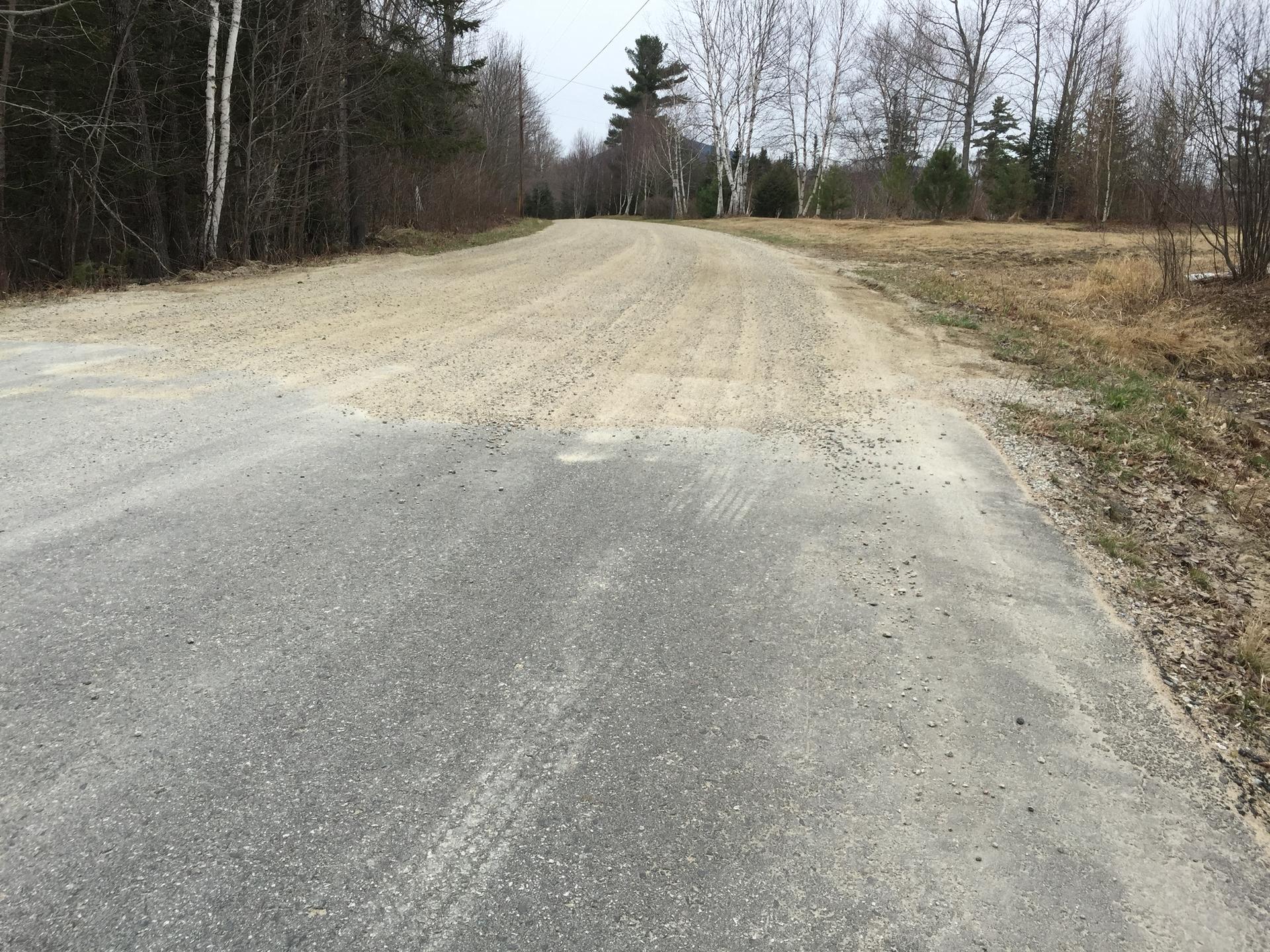 Gravel road meets paved road in a rural, overcast landscape.