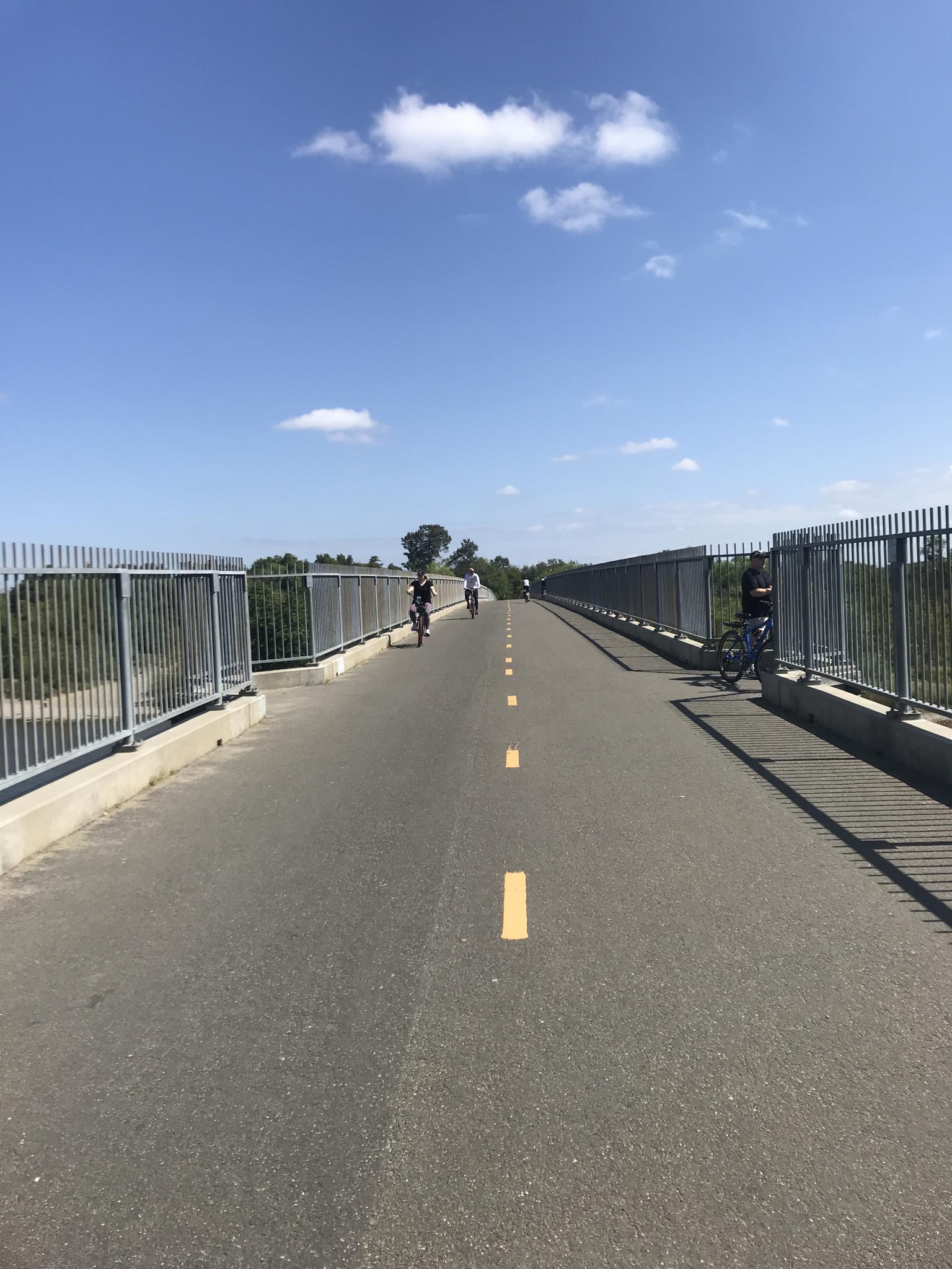 Sunny day on a fenced paved path with people cycling in the distance.