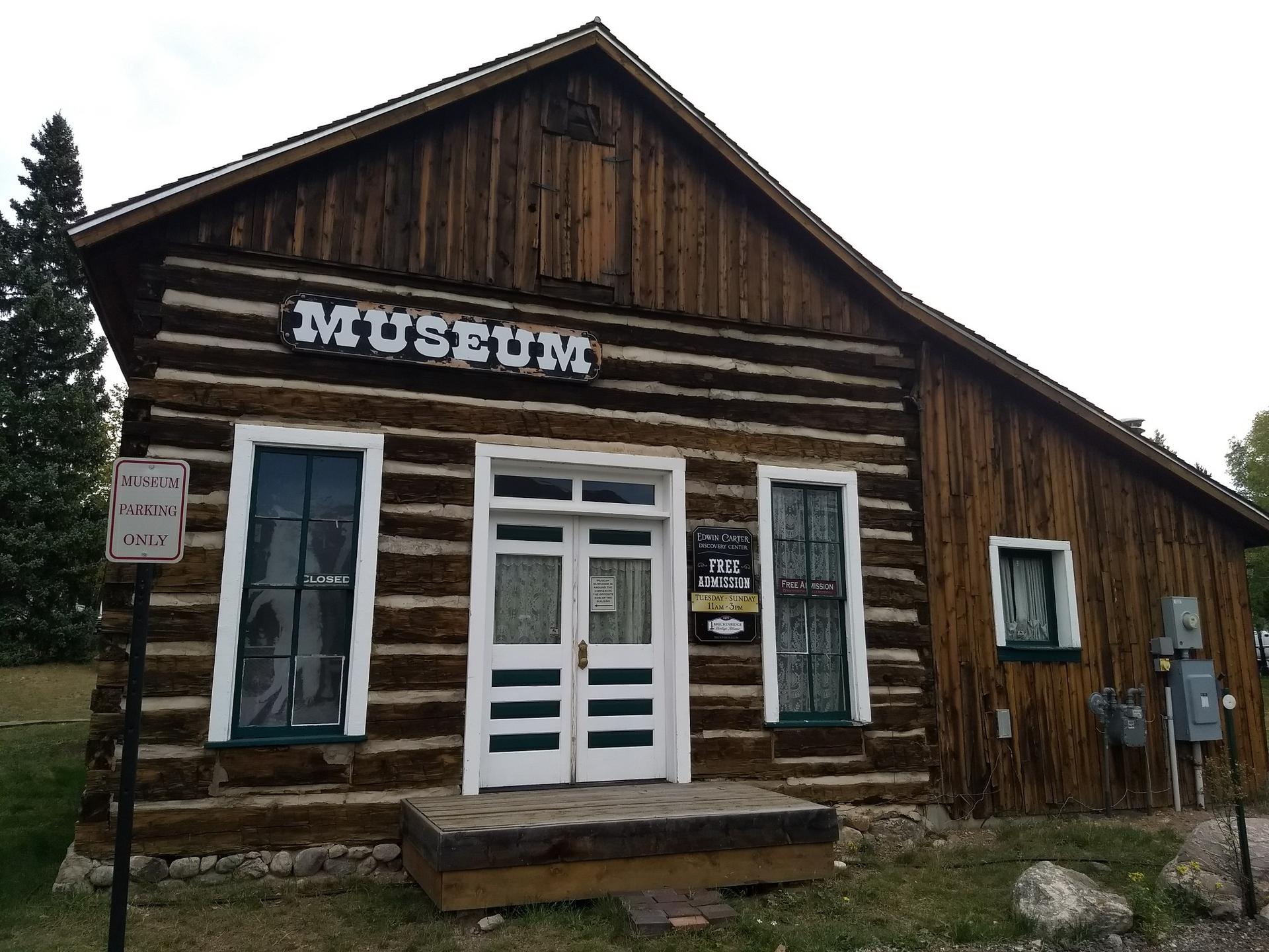 Rustic log cabin museum building with white double doors under an overcast sky.