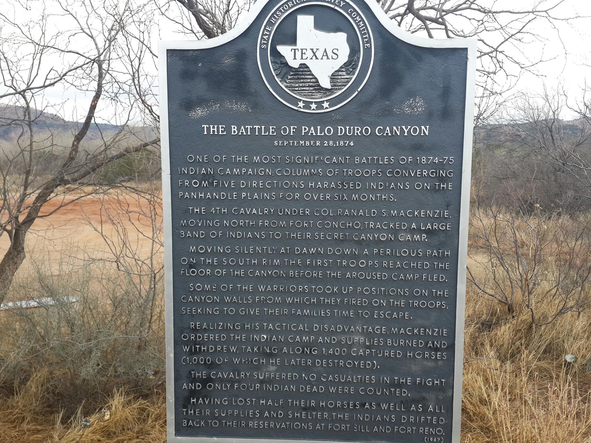 Black historical marker with white text, surrounded by dry brush.