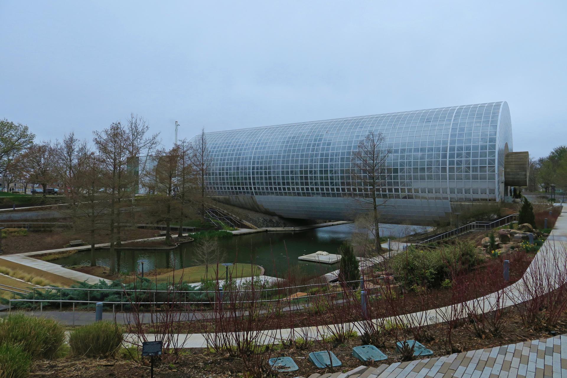 Modern glass building beside a pond and gardens on an overcast day.