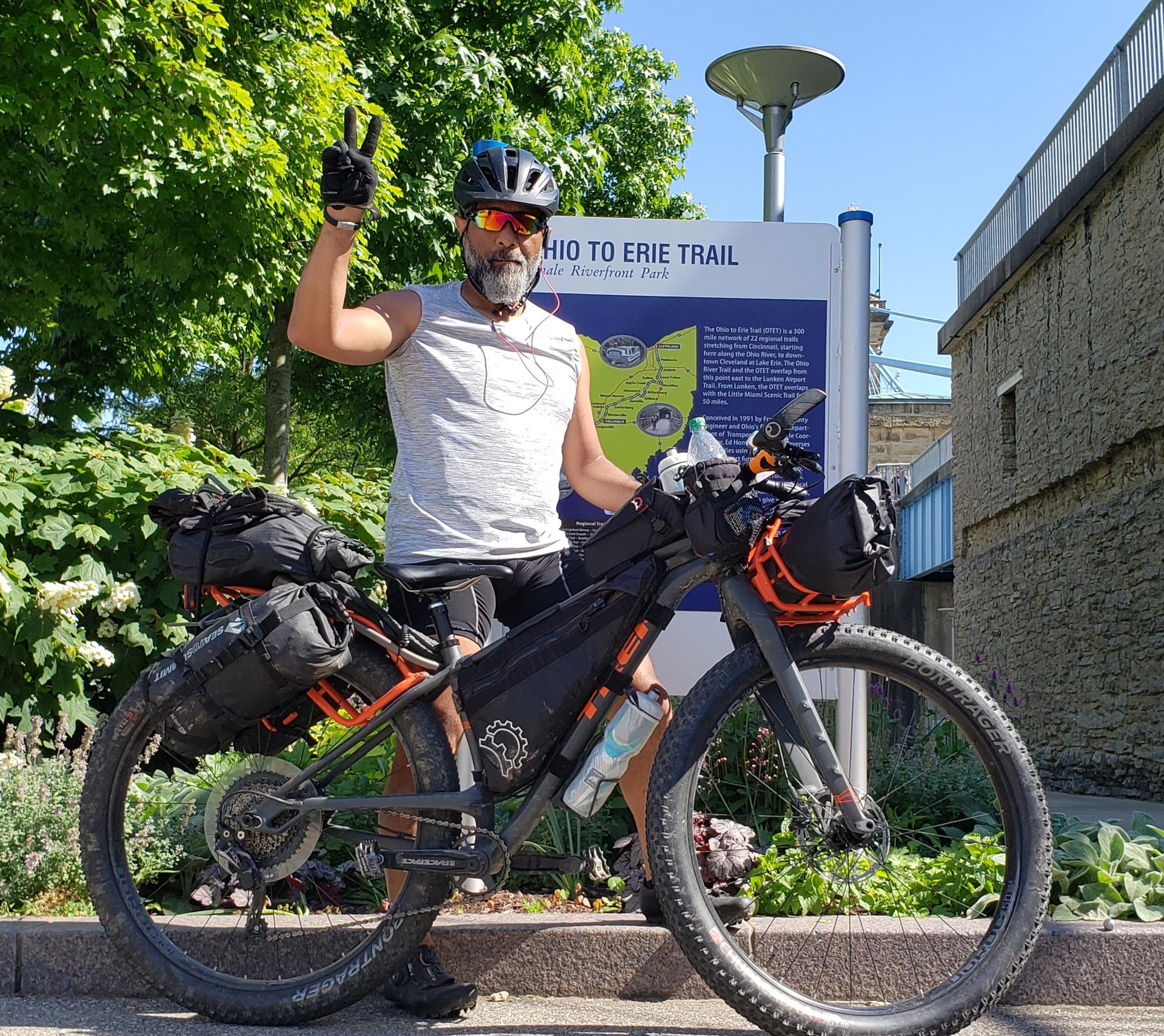 Smiling man on a loaded touring bicycle gives a peace sign. Bright daylight.