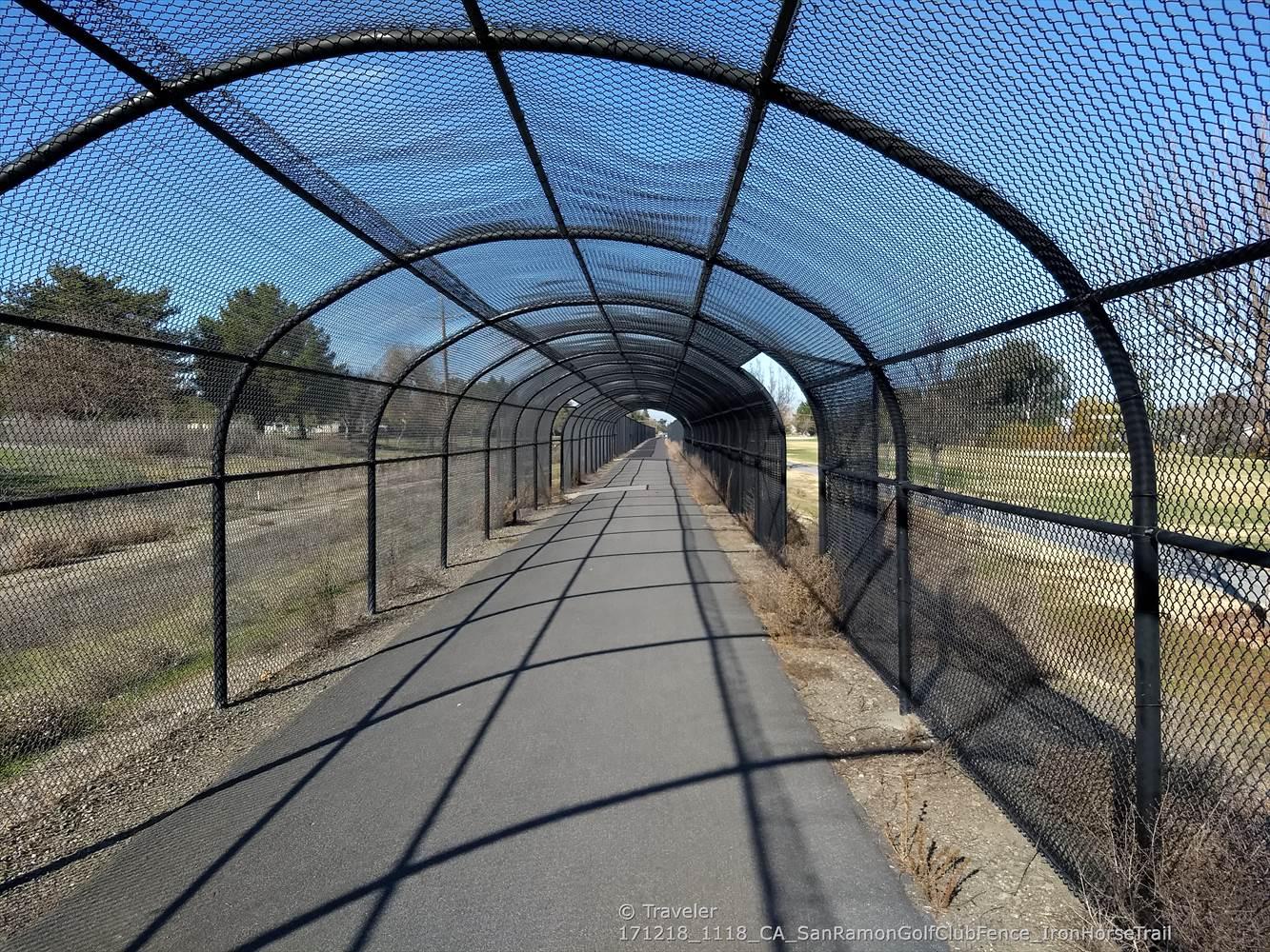 Long, arched mesh tunnel covers a paved path on a bright sunny day.