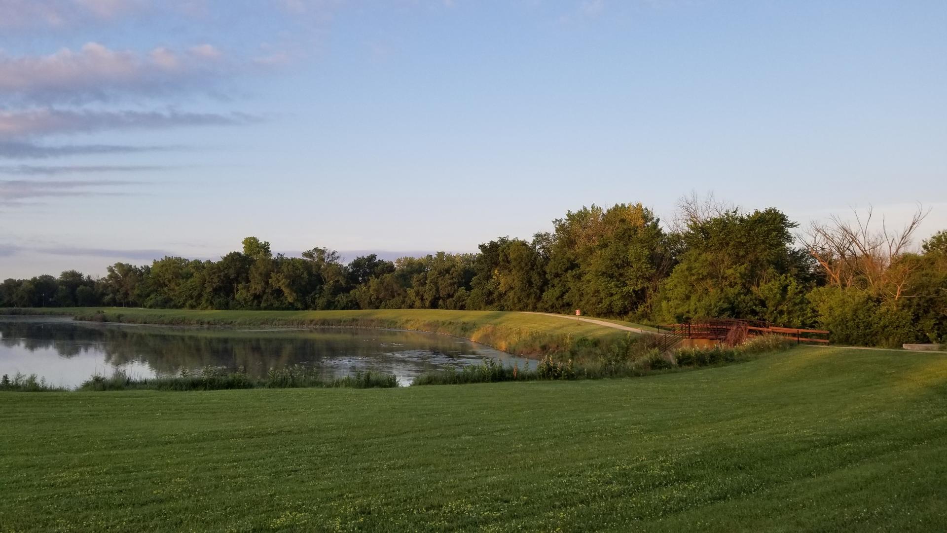 A calm lake reflects trees, with a grassy foreground and distant path.