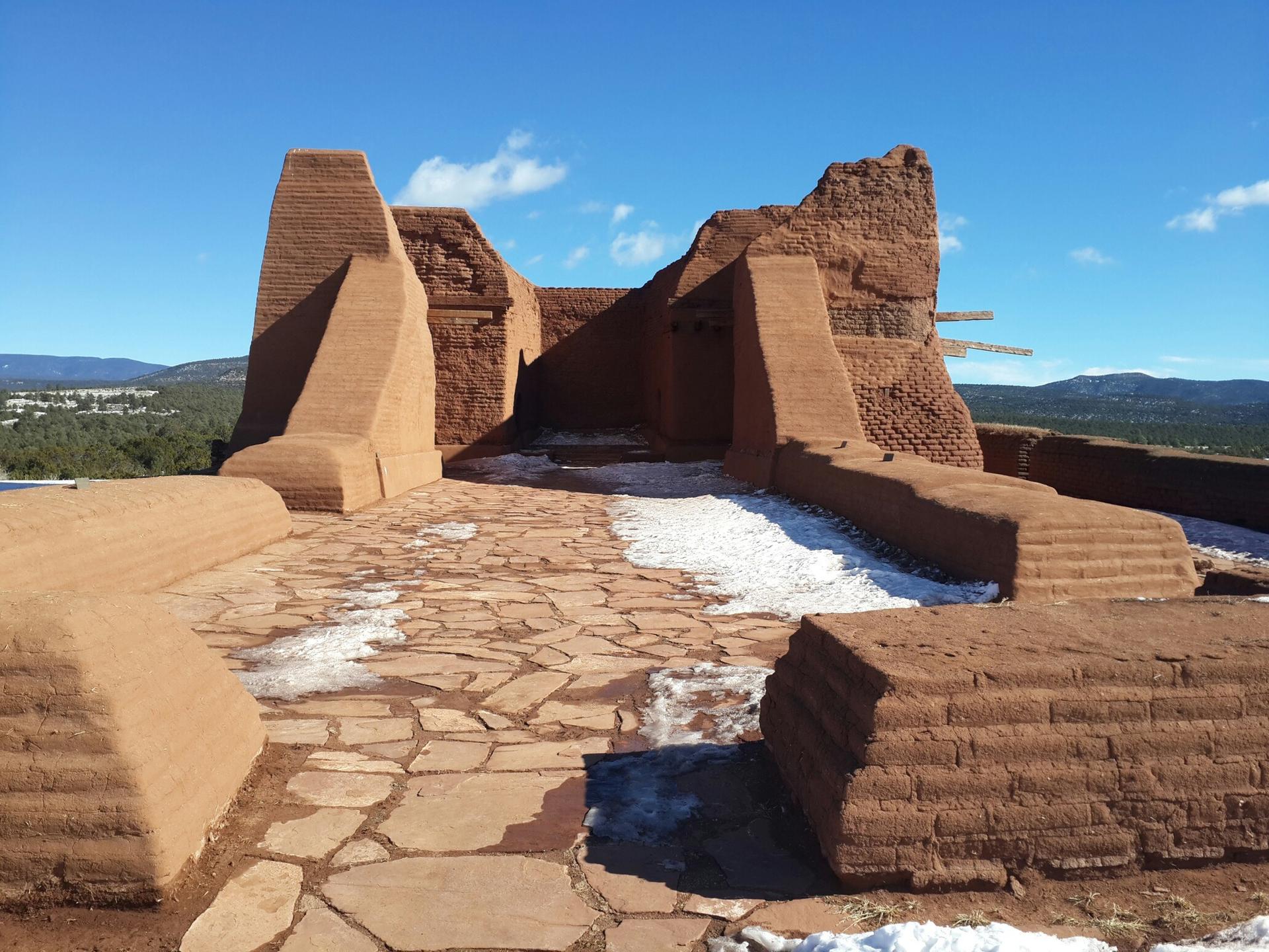 Large adobe-style structure with stone path and snow patches under blue sky.