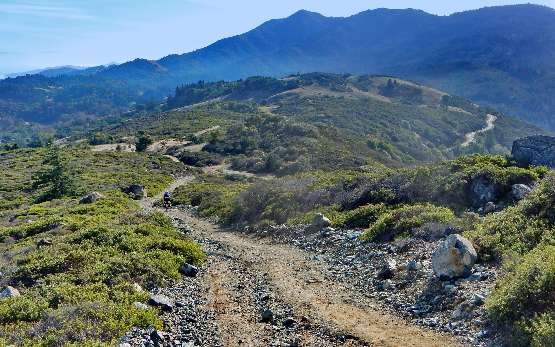 A winding dirt trail through green hills with distant mountains under a clear sky. A person bikes.