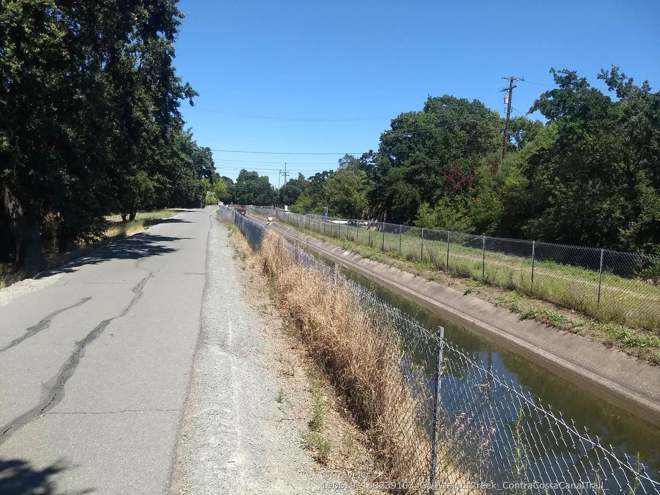 Paved road alongside a fenced canal, with green trees under clear sky.