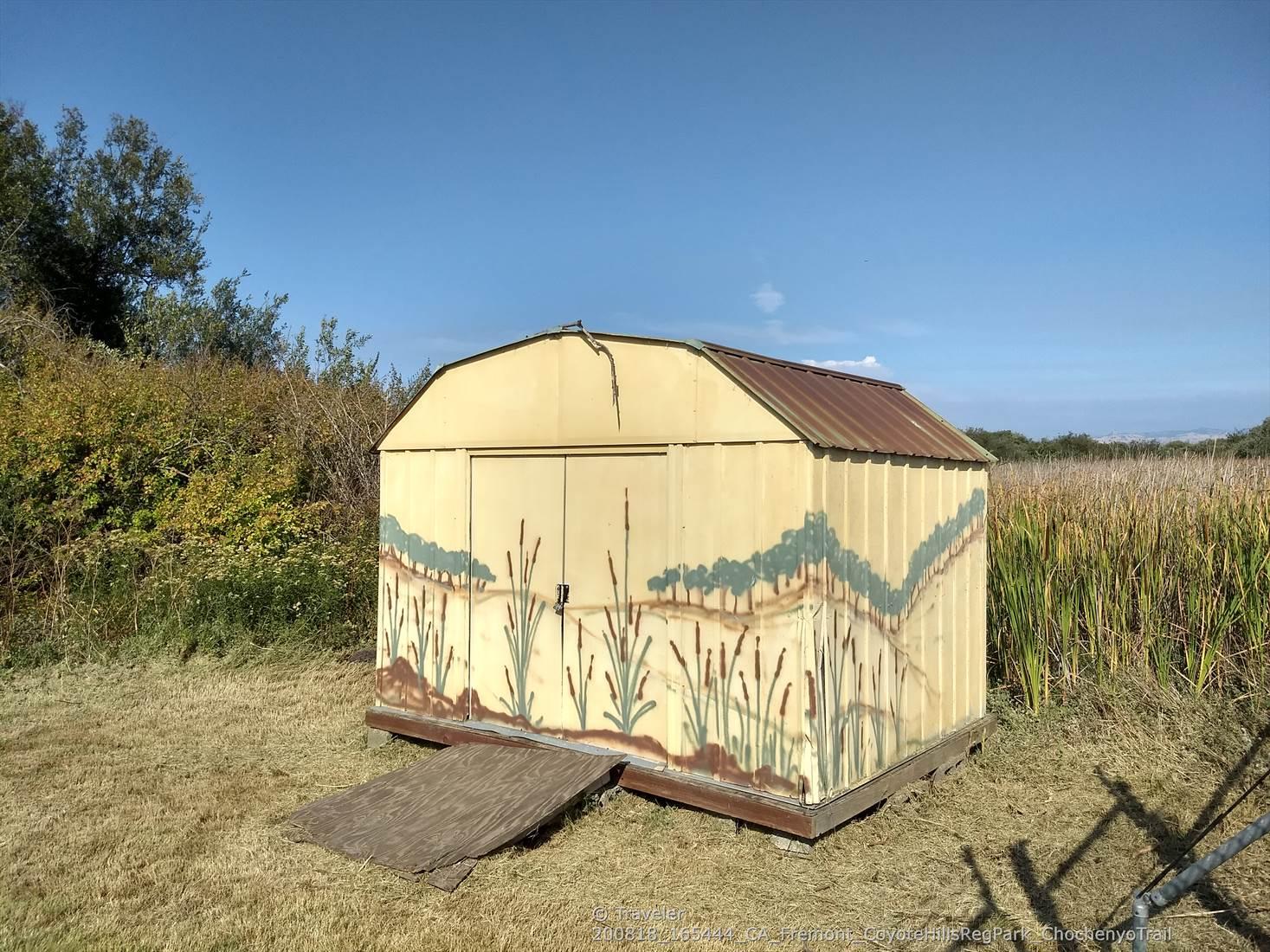 Small shed with painted reeds and hills, surrounded by dry grass.