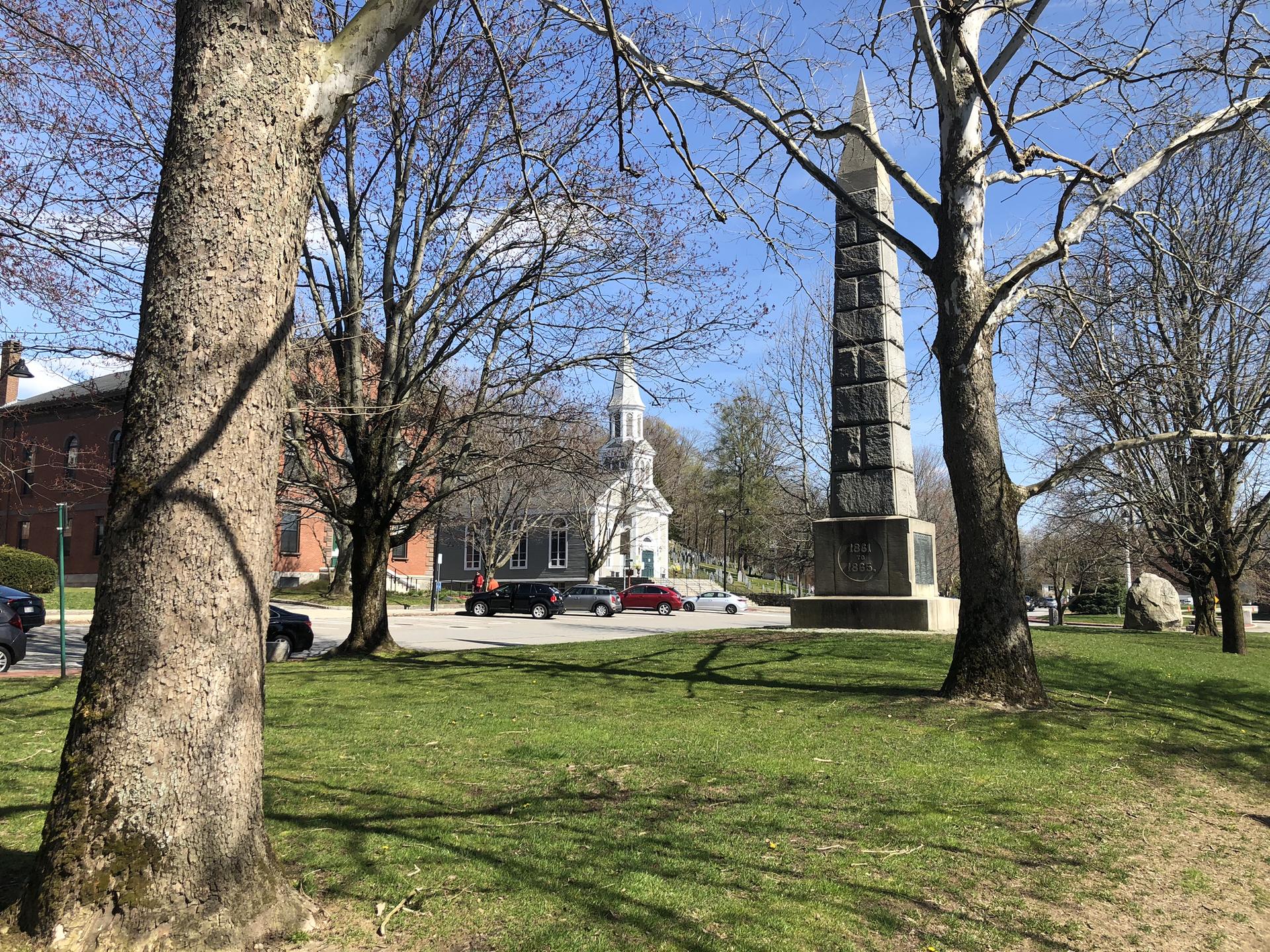 A park with large trees, a tall monument, and a distant white church.