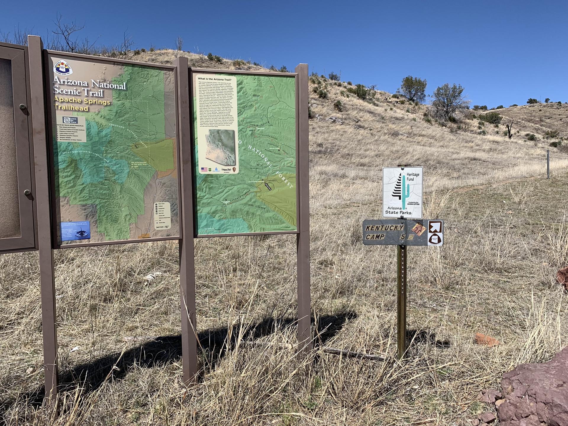 Information boards and a trail sign on a dry grassy hillside in daylight.
