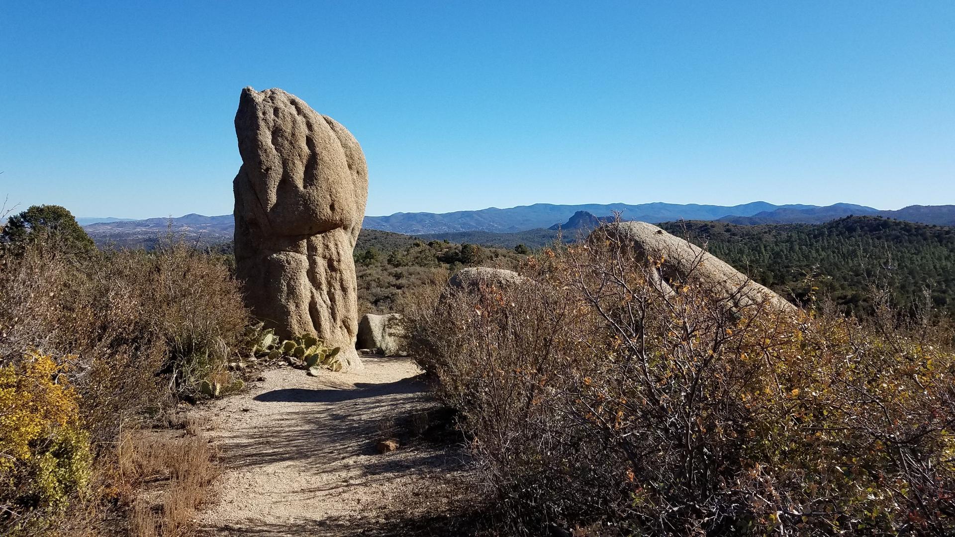 Tall, weathered rock formation on a dirt path under a clear blue sky.