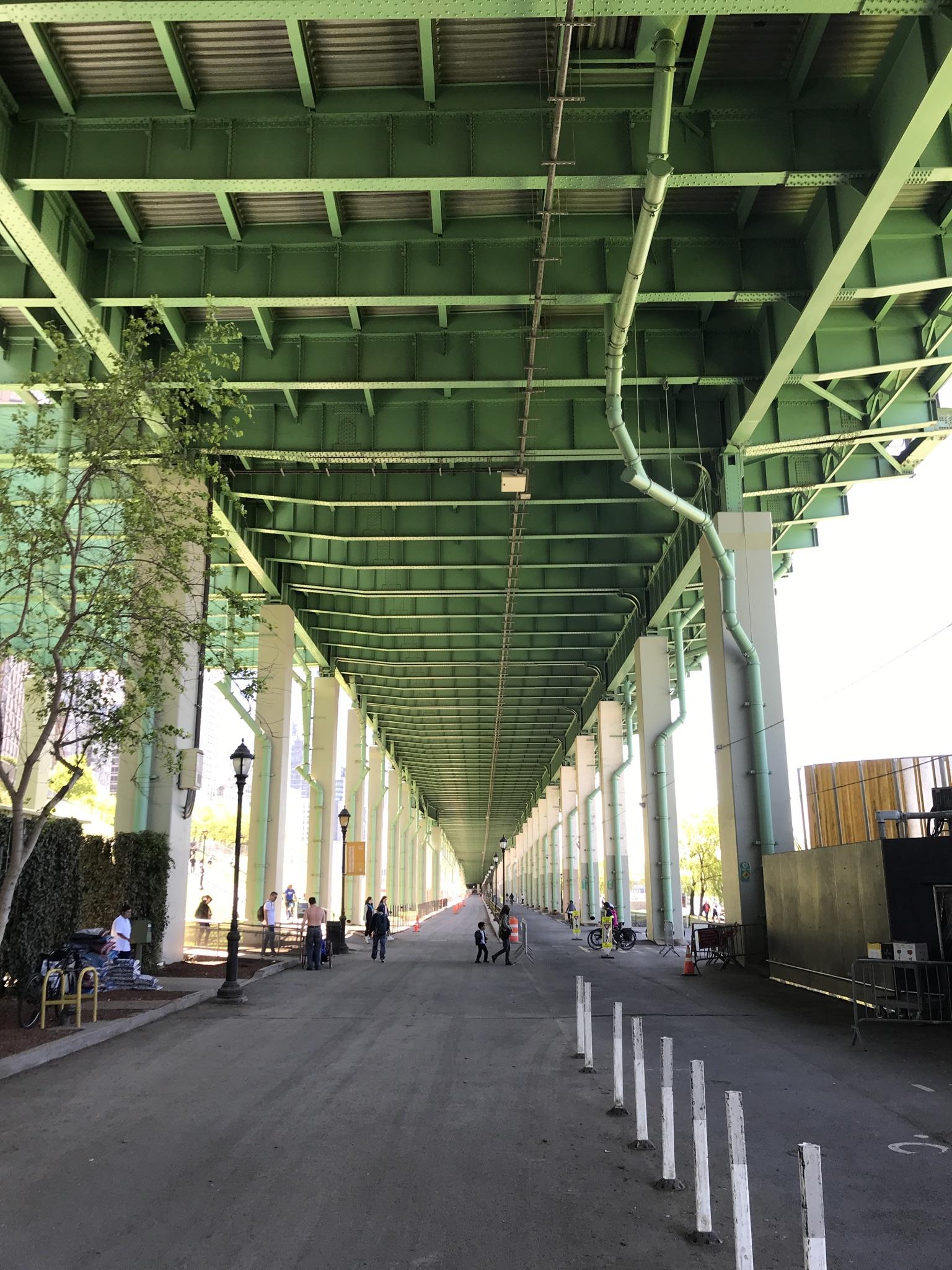 People walk under a long, green, elevated bridge structure on a bright day.