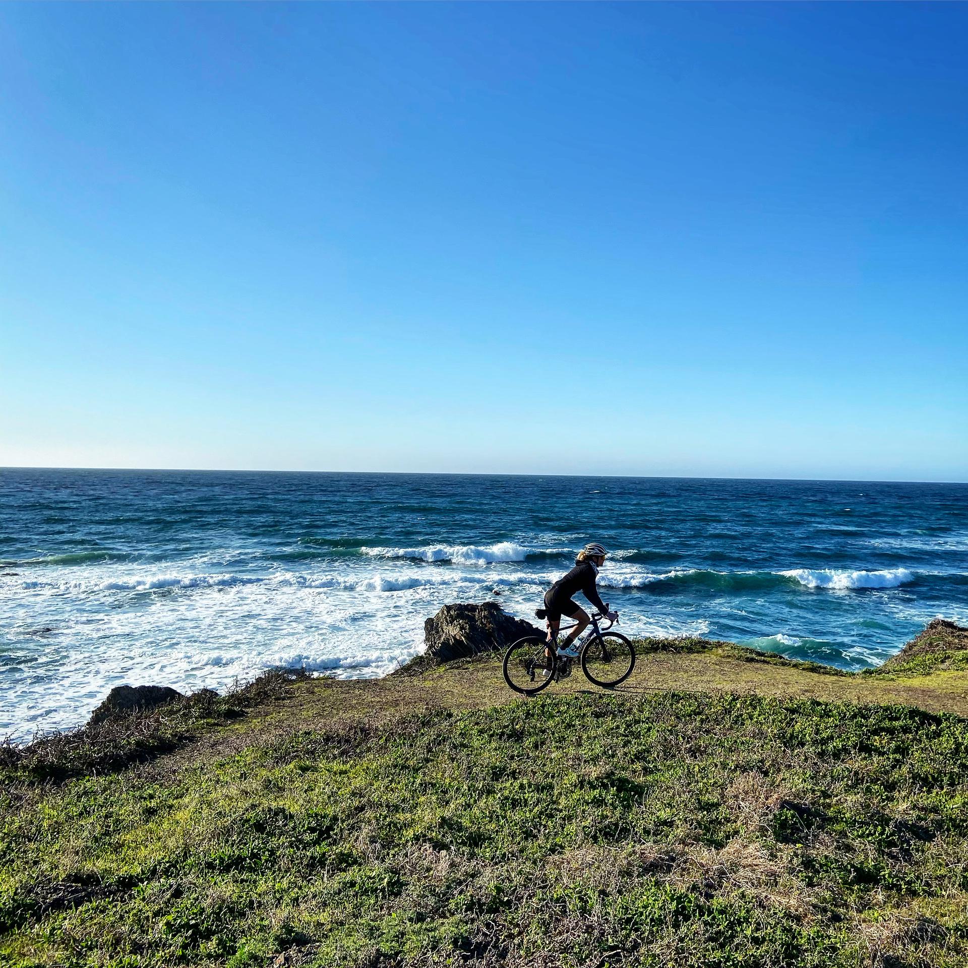 A person cycles on a grassy cliff overlooking a wavy blue ocean.