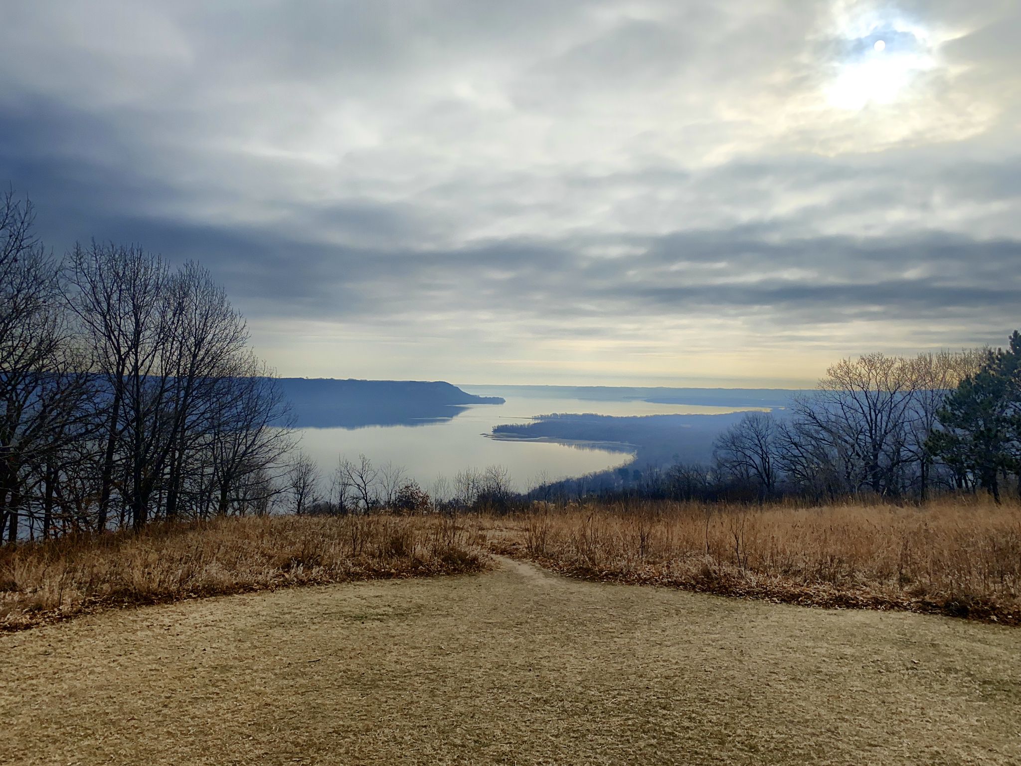 Panoramic view of a wide river valley with cloudy skies and sun peeking through. - Bluffside Trail