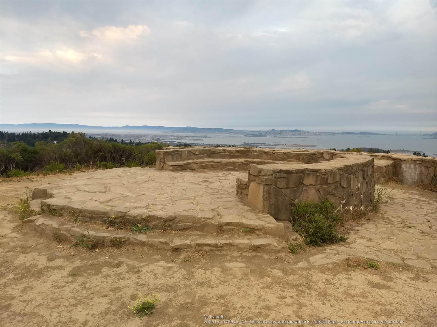 Circular stone structure on a hilltop overlooking a bay under cloudy skies.