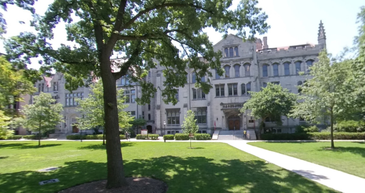 Grand stone building behind a sunny green lawn with a large tree.