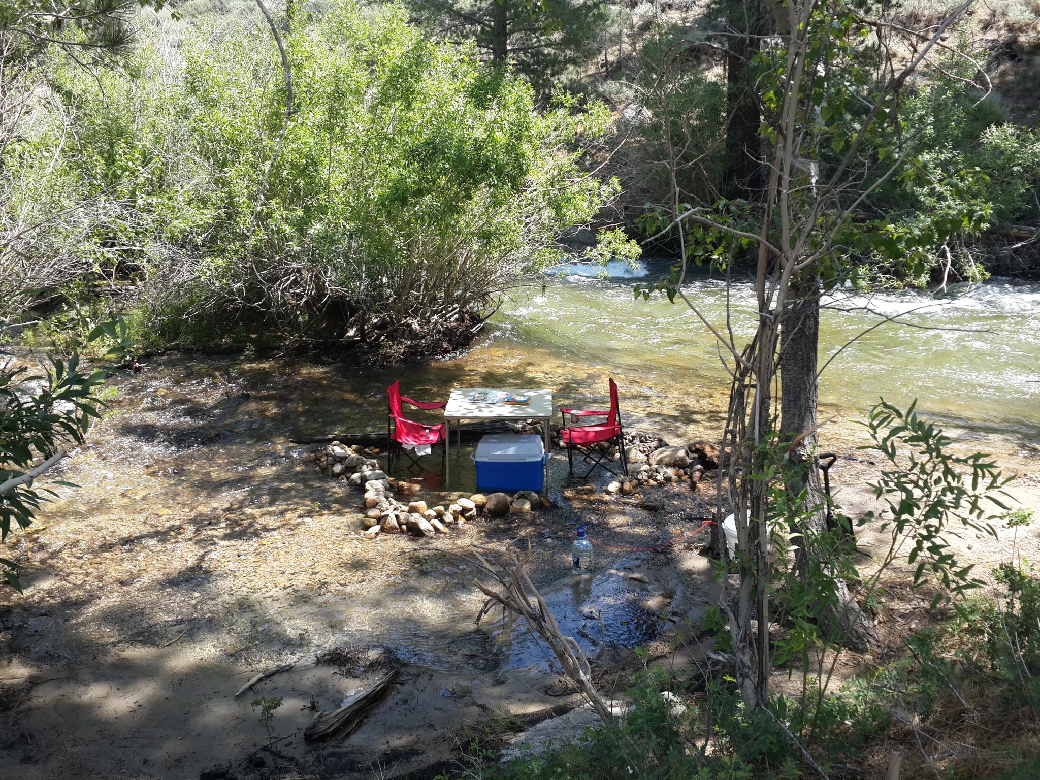Two red chairs and a table by a flowing river in a sunny, wooded area.