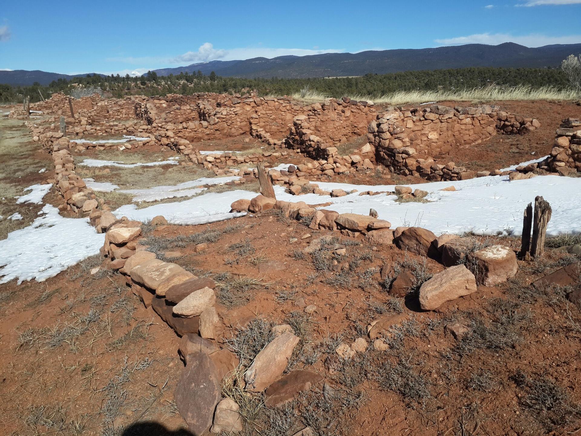 Ancient stone ruins on arid ground with patches of snow under clear sky.