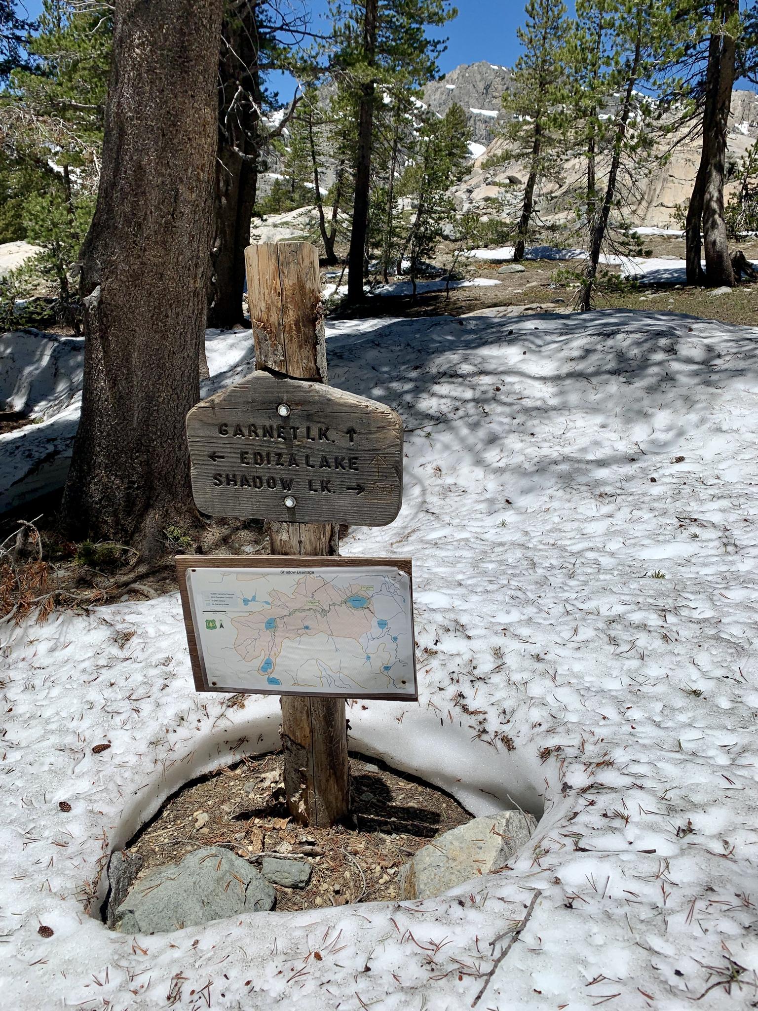Wooden trail sign and map surrounded by snow in a sunny forest.