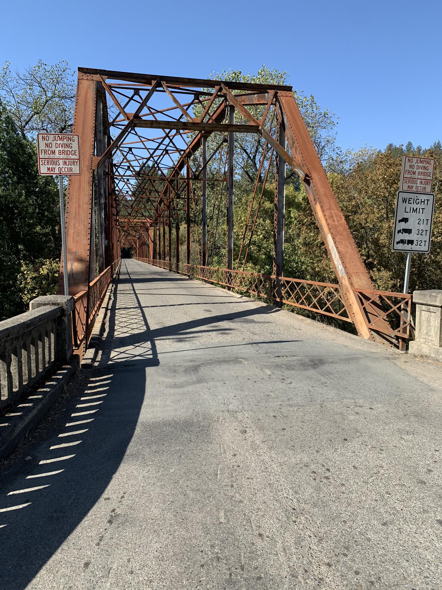 A rusty metal truss bridge spans a road, bathed in bright sunlight.