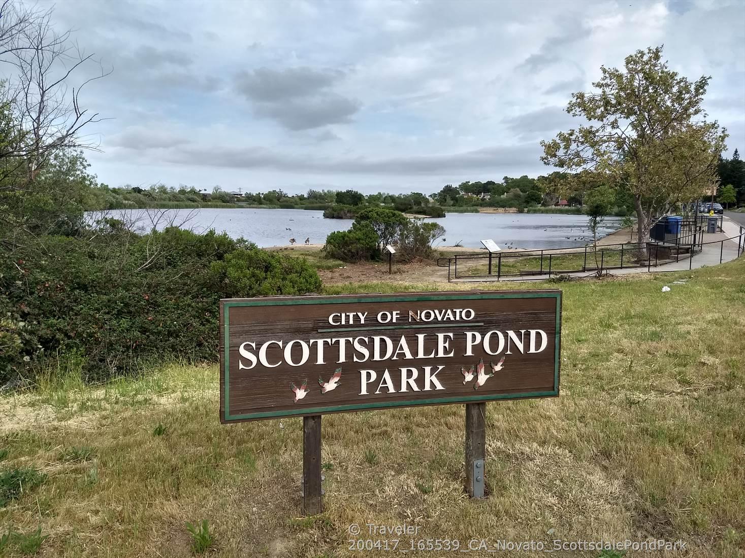 A wooden park sign in front of a large pond under a cloudy sky.