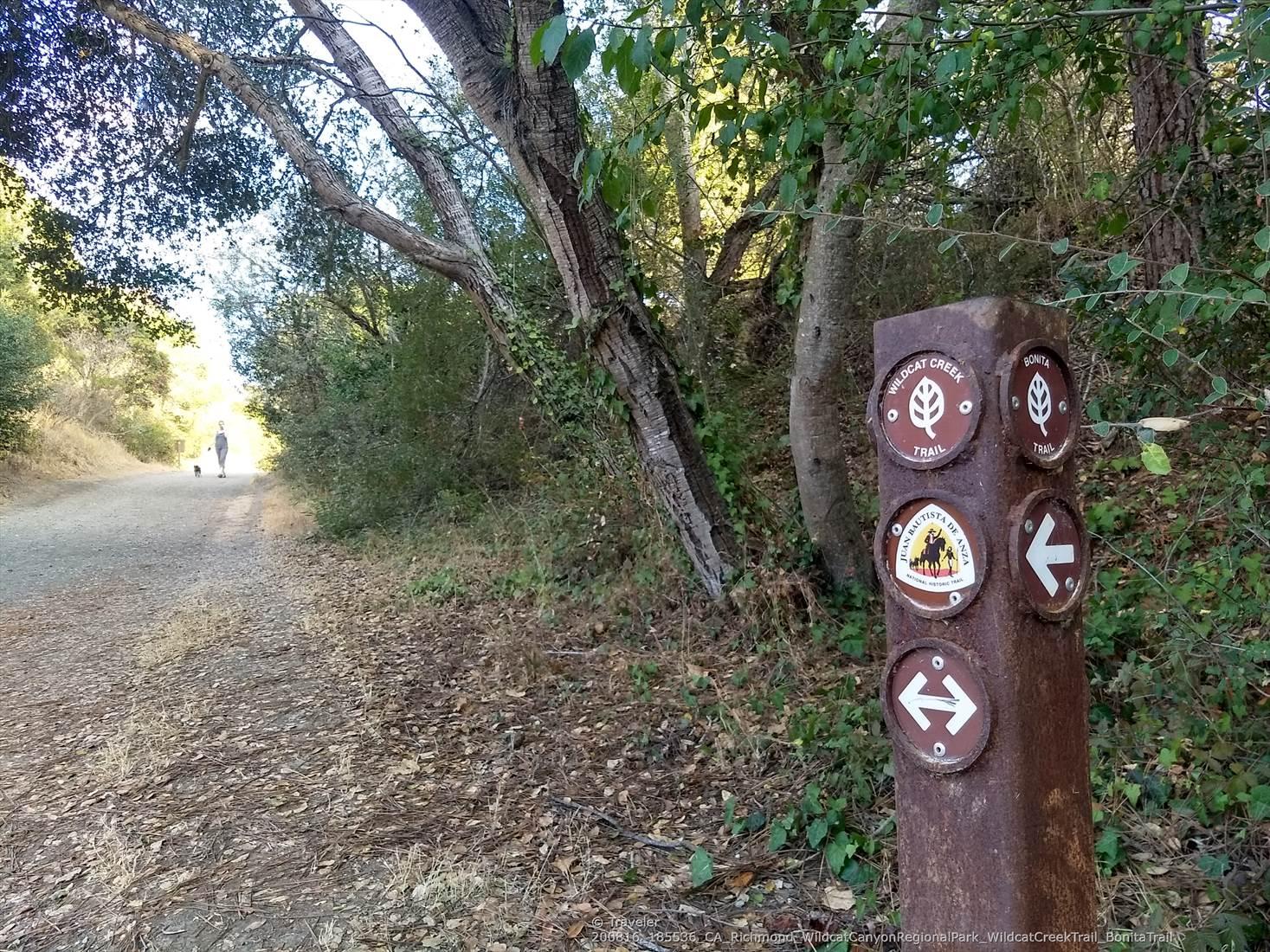 A trail marker post beside a dirt path, a person walks in the distance.