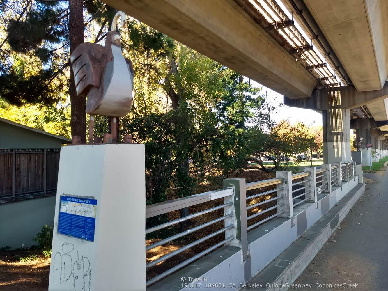 A large bird sculpture stands under an elevated concrete pathway on a sunny day.