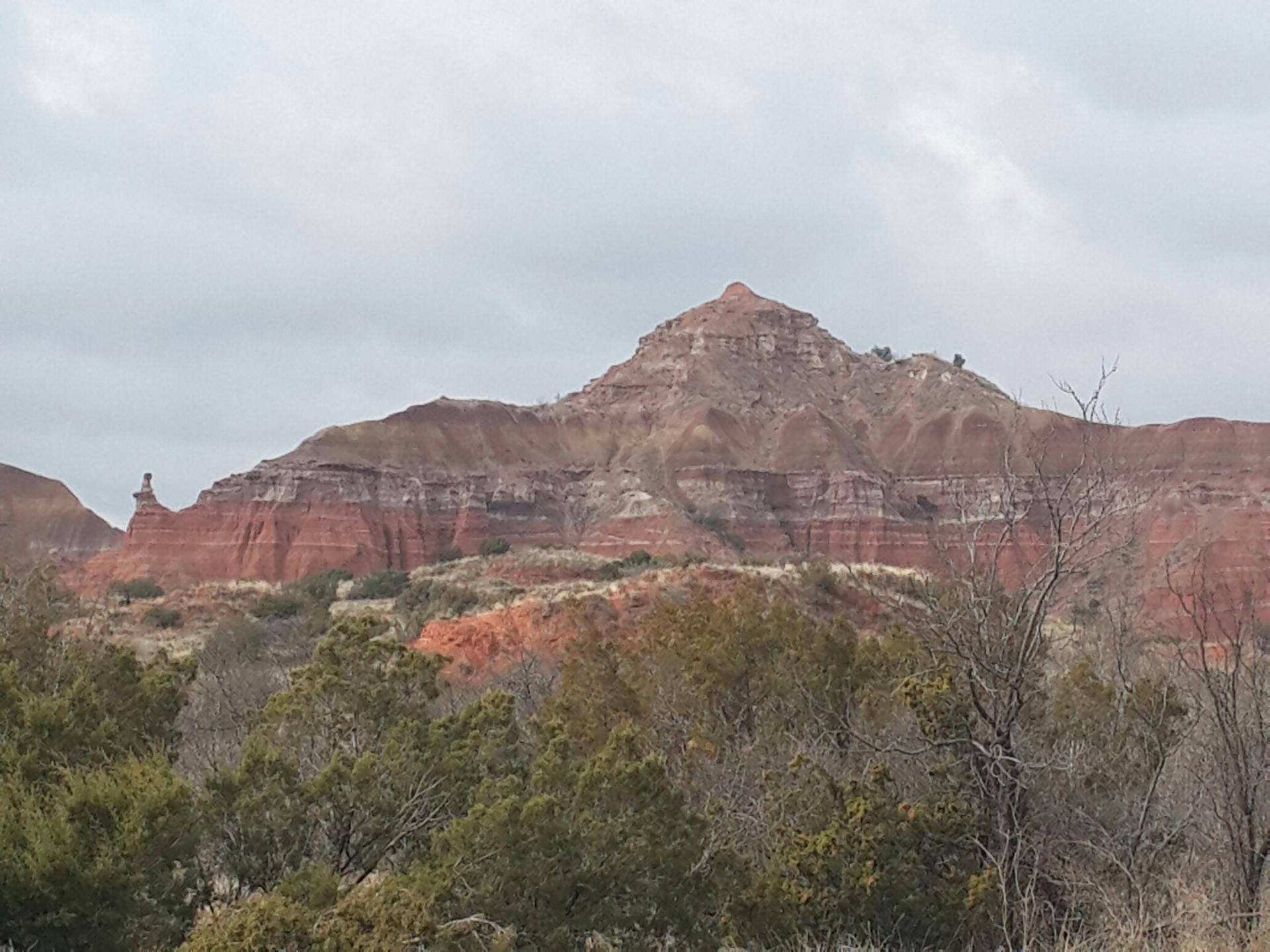 Red and white layered mountains with green shrubs under a cloudy sky.