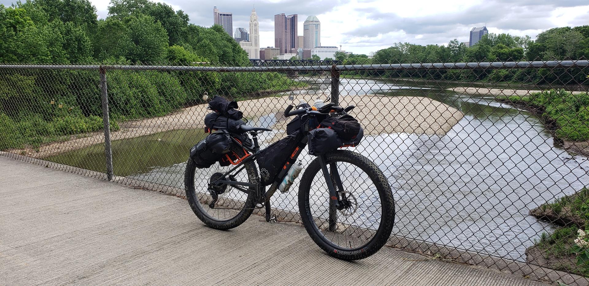 Bikepacking bicycle parked on a bridge next to a river and distant city. Overcast.