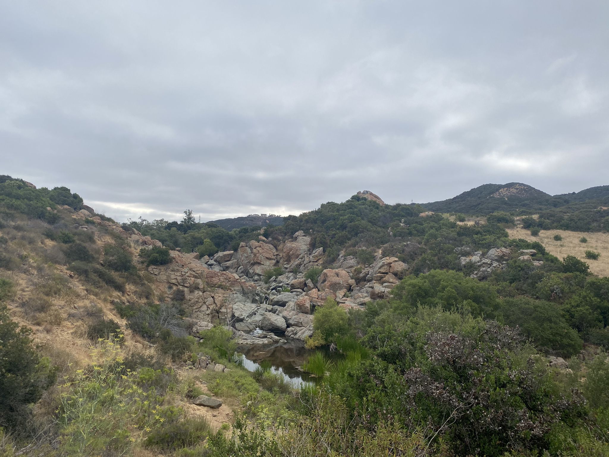 Overcast view of a rocky canyon with a stream and green hills. - Los Penasquitos Canyon Trail