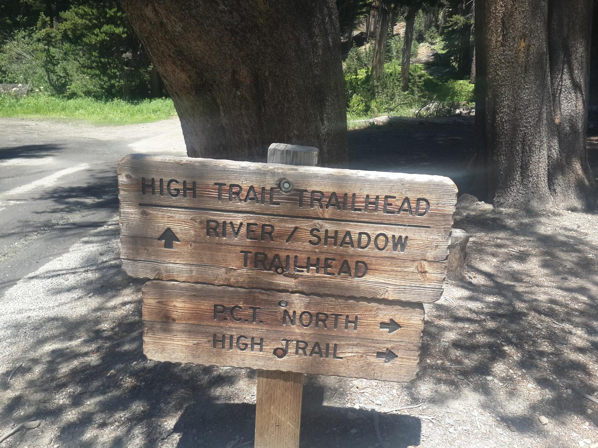 A wooden trail sign with directional arrows, partially shaded by trees.