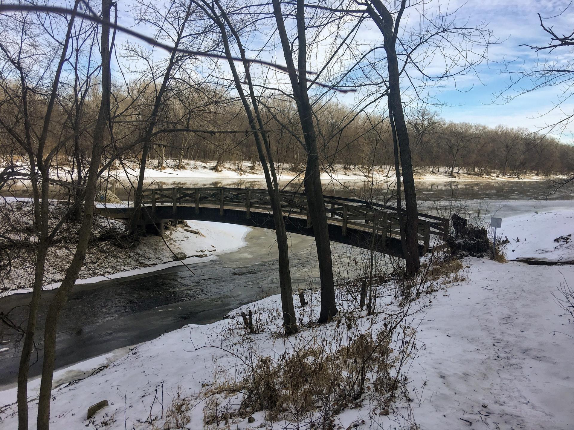 Wooden bridge over a partially frozen river with snow-covered banks and bare trees.